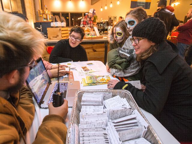 Three young people sit at a table at an event with an open laptop and seed packets in front of them talking to two young girls with their faces painted to look like skeletons.