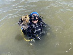 A person wearing googles and a black wet suit holds up a big oyster.