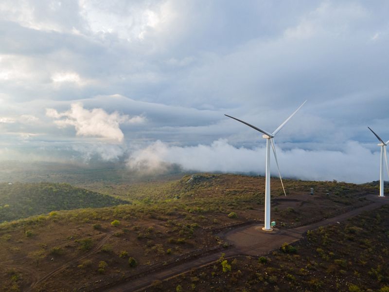 View looking across a mountainous expanse, with two large wind turbines in the foreground.