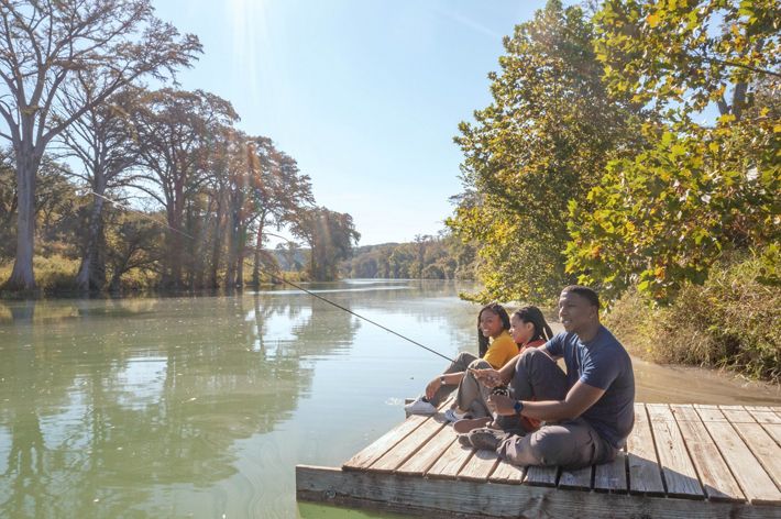 Three people sit on a dock fishing on a turquoise river lined with trees.