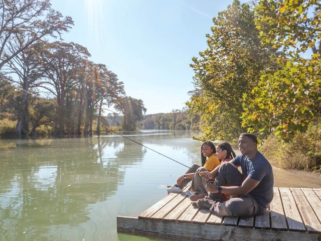 Three people sit on a dock fishing on a turquoise river lined with trees.