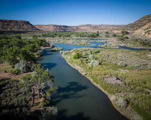 A wide river is flowing on a landscape with green shrubs and mesas in the background.