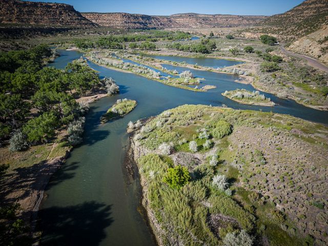 A wide river is flowing through a landscape with green shrubs and mesas in the background.