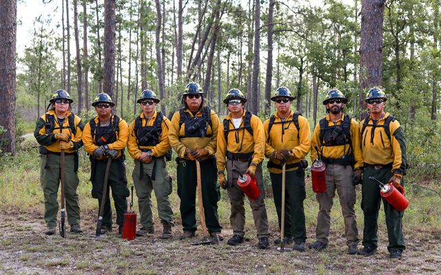 Eight members of the Alabama-Coushatta Tribe of Texas pose together in yellow protective fire gear, holding drip torches and axes.