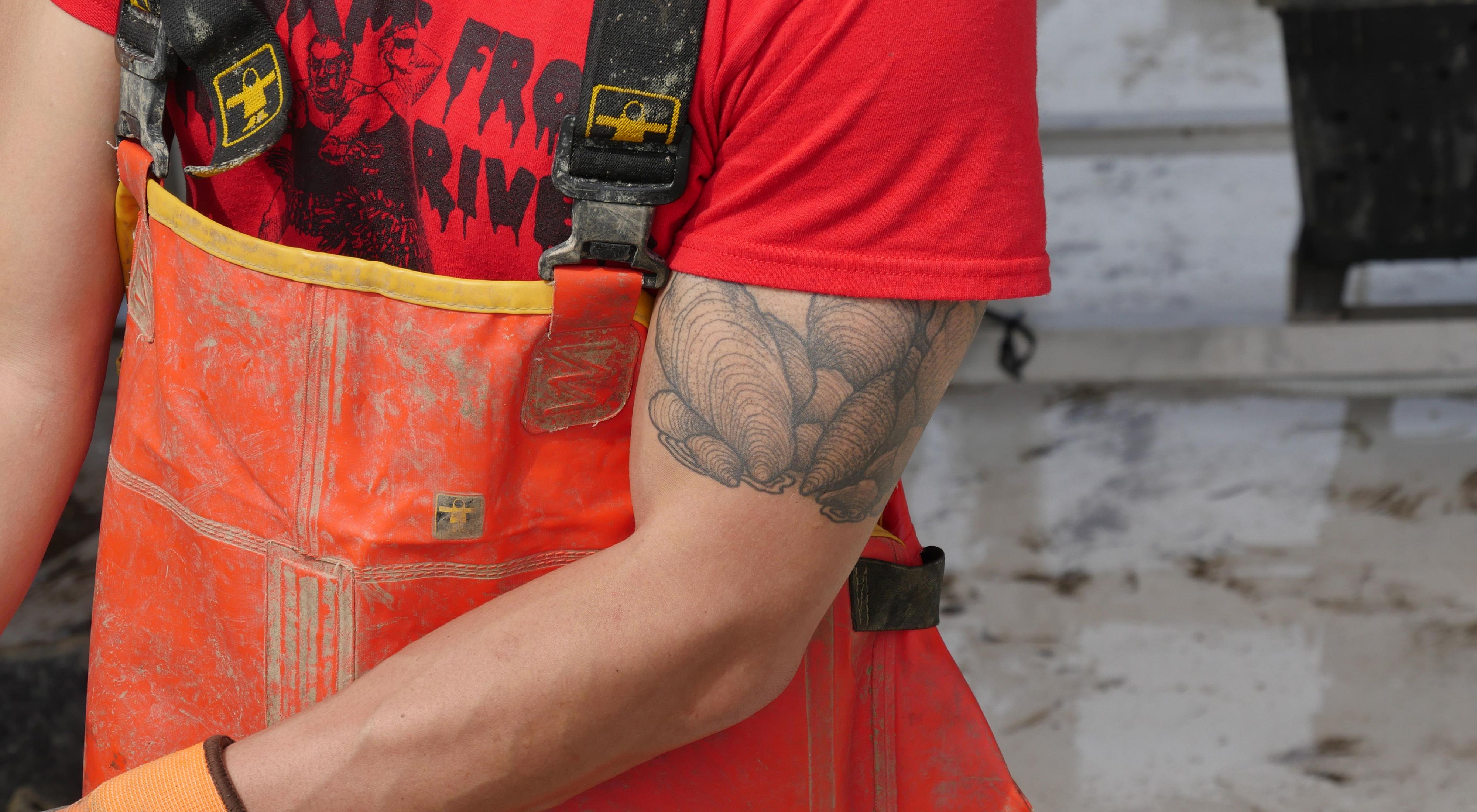 Close cropped view of a man man wearing orange chest waders shucking oysters. He has a large tattoo of a clump of mature oysters on his bicep visible beneath the sleeve of his red tshirt.