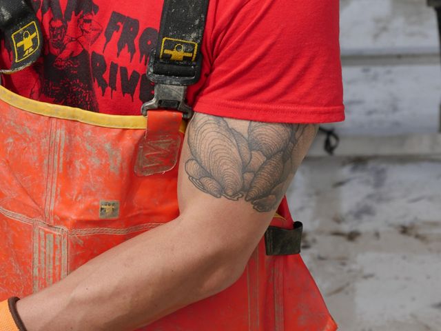 Cropped view of a man shucking an oyster. He is wearing orange overalls. A large oyster tattoo is visible on his bicep.