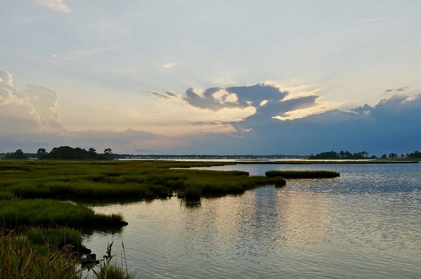 The sun sets over coastal wetlands in Maryland. Slim fingers of marshy land extend into the wide, rippling water. The setting sun illuminates a large bank of clouds gathering on the horizon.