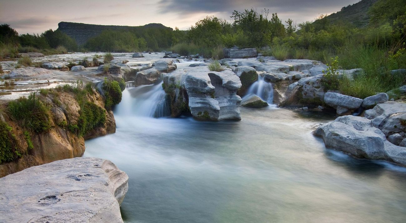 Turquoise waters flow and fall over stacked rocks surrounded by greenery.
