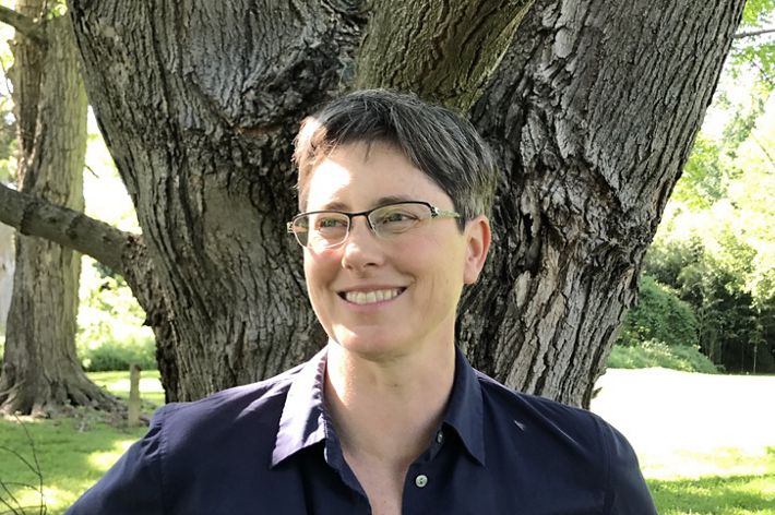 Donnelle Keech headshot. A smiling woman wearing a dark blue shirt stands in front of tree. A bright patch of sun is visible in the grass behind her.