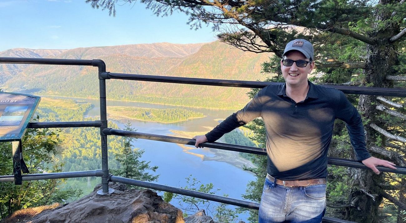 A smiling man poses in front of a valley overlook. A wide river curves along the valley floor at the base of a tree covered mountain.