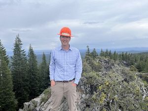 A man wearing an orange hard hat stands at the edge of a rocky outcrop. The tops of tall evergreens are visible in the background, growing up from the valley below.