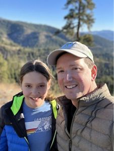 A man and girl pose together in front of a rolling mountain ridge.