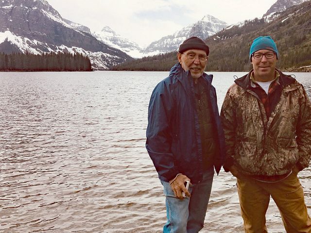 Two men in pose in front of a mountain lake.