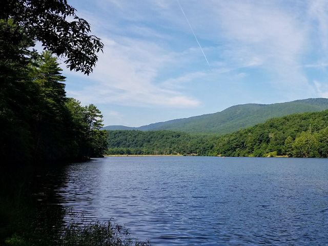 A wide blue lake is ringed with mature trees. A low dam is visible at the far end. A mountain ridge rises behind the lake.
