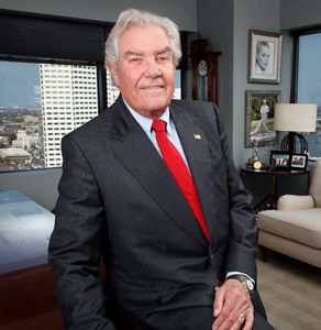 A man wearing a dark suit and red tie smiles at the camera.
