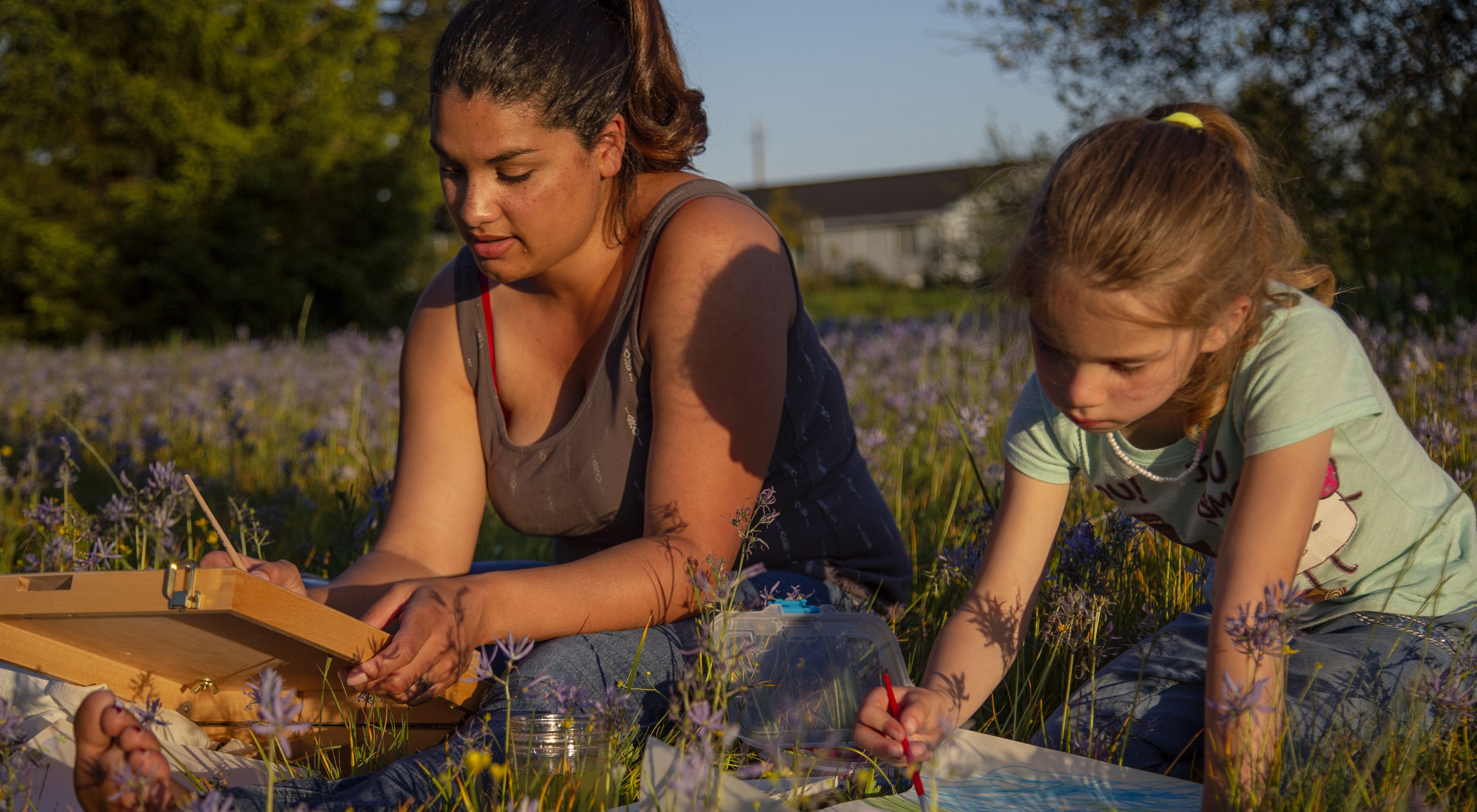 Kenison sits beside daughter-hair in ponytail-in a field of high purple meadow flowers, each with a canvas and paintbrush. Both focused on painting. House sits in distance.