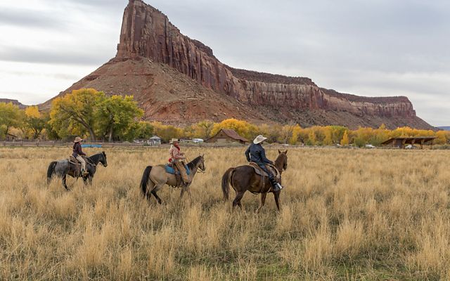 Three people in jeans and western hats ride horses in high grass. A red rock formation juts sharply upward in the distance.