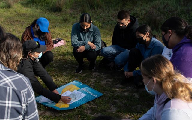 Students and an environmental educator sit in a circle on the ground looking at a map of the region. 