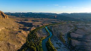 A blue river lined with brush winds through arid cliffs.