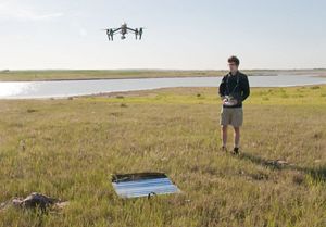 A young person stands in a field and flies a drone in front of him.