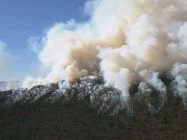 Large clouds of white smoke lift off of a barren hill into a blue sky.