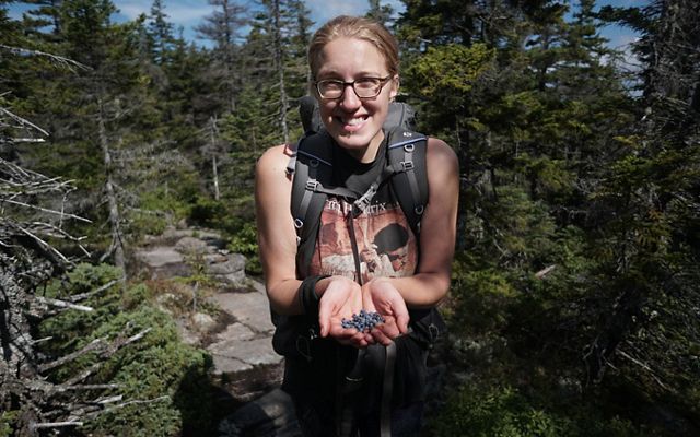 A smiling woman holds freshly picked wild blueberries in her cupped hands. She is standing on a trail surrounded by tall trees.