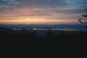 The sun hangs low on the horizon at sunset streaking the sky pink and orange. Darkly shadowed evergreen trees frame the foreground.