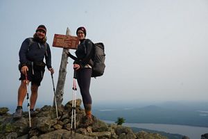 A man and woman stand in front of a sign marking the peak of Bigelow Mountain. The ground is covered with large stones.