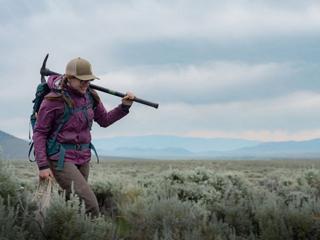 A person carries a pick axe over their shoulder as they walk across a large scrubby landscape.