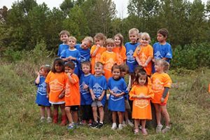 A group of children in orange and blue t-shirts stand in front of a tree-lined riverbank. 
