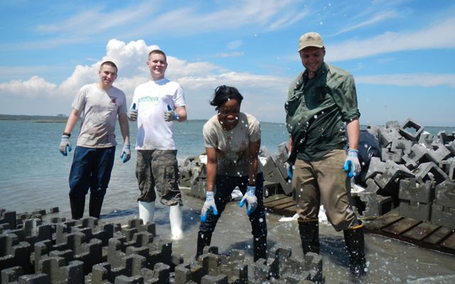 Four volunteers use large interlocking concrete pieces to build oyster castles. Salt spray is kicked up as the waves splash against the blocks.