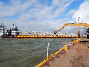An excavator sits on a barge in the ocean, dumping limestone boulders in a bay.