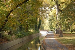 Water rushes over low falls on a narrow creek. A paved path runs alongside the water surrounded by trees.