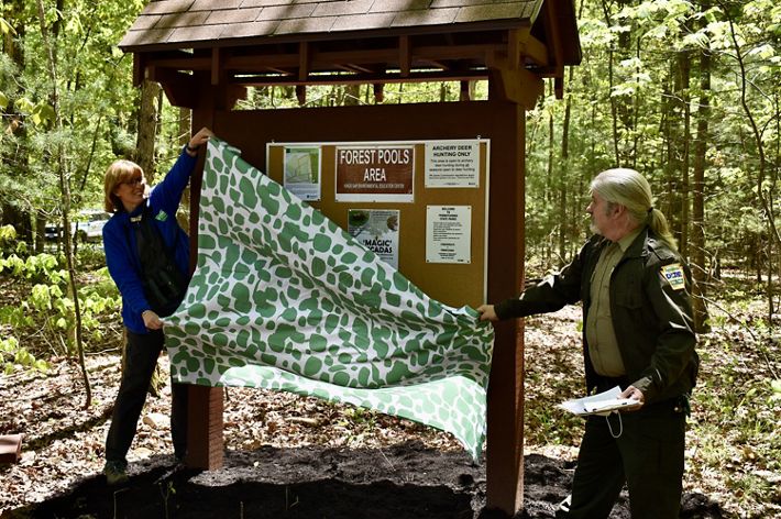 Two people remove a green and white sheet from a new preserve kiosk, part of a land transfer ceremony with the state of Pennsylvania. The kiosk contains signs and visitor information.