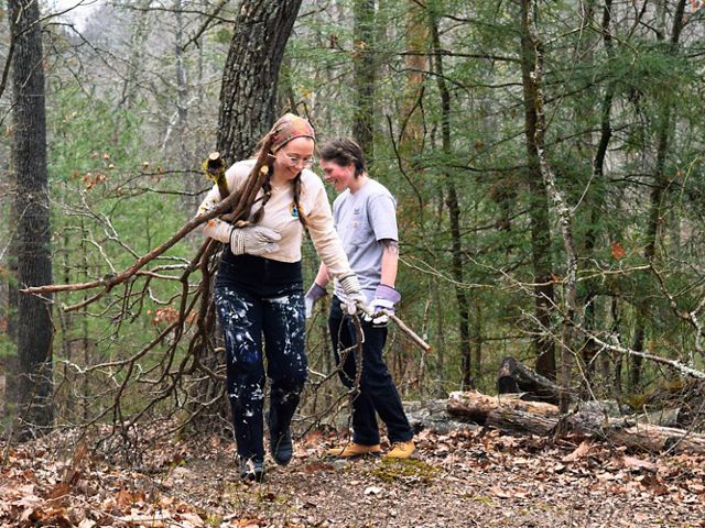 Two people walk in the forest carrying cut branches and vegeation.
