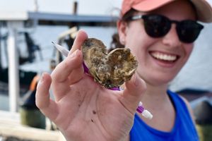 an oyster shaped like a heart held up by a smiling person in sunglasses and a hat.