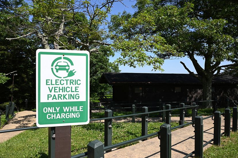 A green and white sign marks an electric vehicle charging station. The sign reads, electric vehicle parking only while charging. In the background, tall trees shade a state park visitor center.