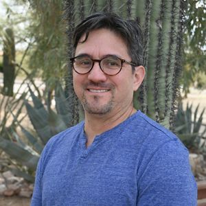 A man in a blue shirt and glasses poses for a headshot in front of a cactus.