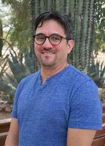 Headshot of a man wearing a blue shirt and glasses smiling at the camera.
