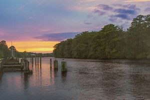 The sun sets over the Pocomoke River, creating bright hues of purple and orange in the sky and reflecting along the water. A wooden dock sits to the right of the river. 
