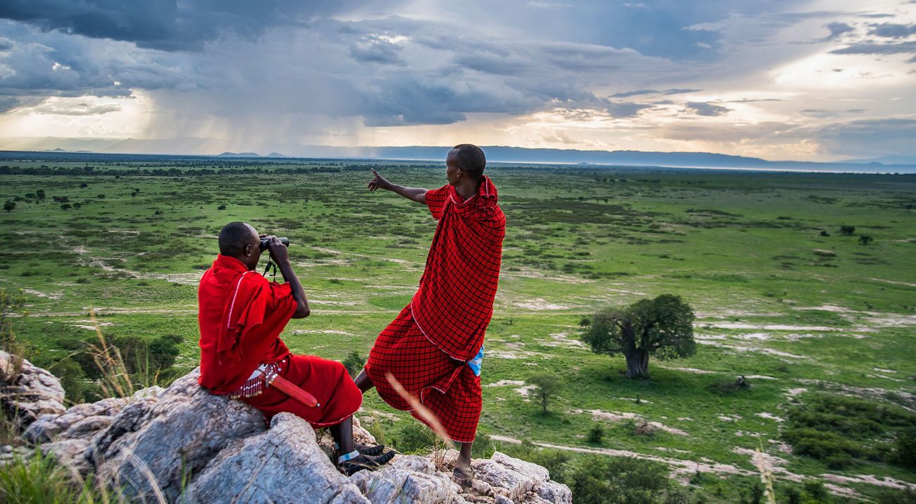 Two people in bright red traditional fabrics look out over a wide grassland, pointing at a rainstorm in the distance.