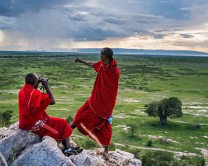 Two people in bright red traditional fabrics look out over a wide grassland, pointing at a rainstorm in the distance.