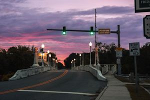 A paved roadway stretches into the distance over the Pocomoke River, beneath a vibrant sunset sky painted in shades of pink and purple.
