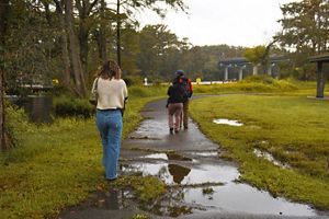 Three people walk along a path near a river. It has just rained and there is evidence of flooding on the path. 
