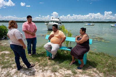 Two people sit on a bench and two people stand talking to them at the edge of an ocean.