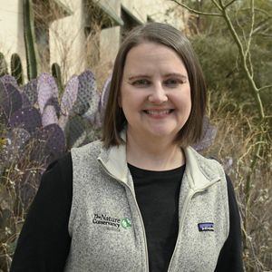 A woman in a grey vest smiles for a headshot with a purple cactus behind her.