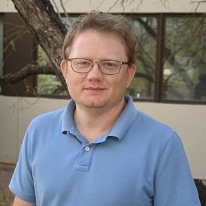 A man in a blue shirt and glasses poses for a headshot in front of a building.
