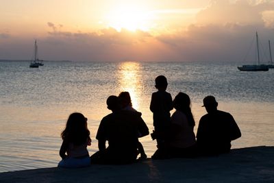 A family of several adults and children are silhouetted as they sit on a beach and watch the sun set over the ocean.