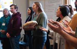 Several people stand in a circle and sing. One of them holds a hand drum.