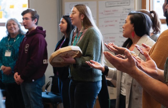 Several people stand in a circle and sing. One of them holds a hand drum.